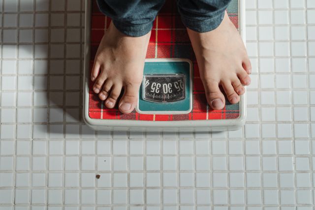 Pessoa medindo o próprio peso em balança analógica. Vista somente dos pés e calça jeans.